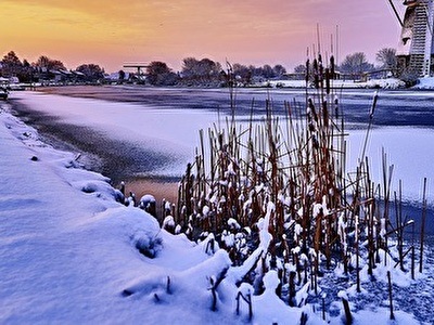 A windmill stands in the snow beside a river, surrounded by a serene winter landscape.