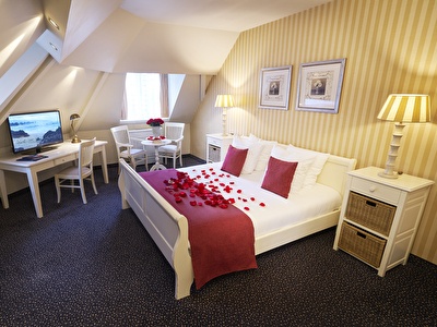 Romantic hotel suite with a double bed, rose petals, a desk with TV, a sitting area and warm lighting.