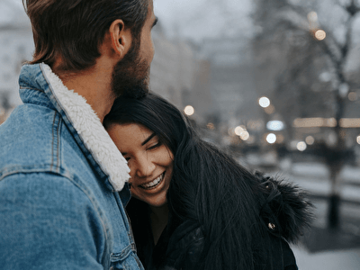 A young couple embracing outdoors in a winter cityscape with soft lights in the background. The woman is smiling as she rests her head on the man, who is wearing a denim jacket with a wool-lined collar.